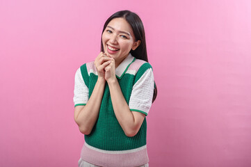 Asian woman smiling sweetly with both hands clasped under her chin, expressing happiness and warmth against a bright pink background