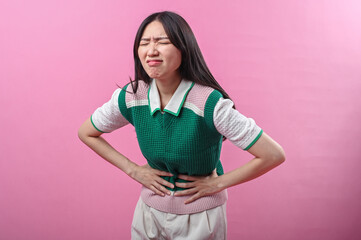 Asian woman clutching her stomach with a pained facial expression, suggesting abdominal discomfort or stomachache, standing in front of a bright pink background