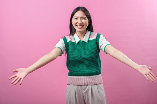 Asian woman smiling warmly with arms wide open as if offering a hug, wearing a green knit vest, standing in front of a pink background