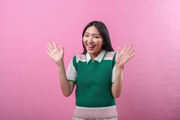Asian woman in green knit top looking surprised and excited with both hands raised near her face, standing in front of a bright pink background