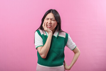 Asian woman holding her cheek with a pained expression, eyes closed in discomfort, possibly experiencing toothache or jaw pain, standing against a plain pink background