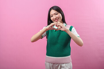 Fototapeta premium Asian woman making a heart shape with both hands while smiling, wearing a green knit vest, standing against a solid pink background