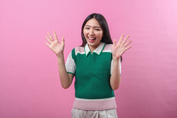 Asian woman in green knit top looking surprised and excited with both hands raised near her face,...
