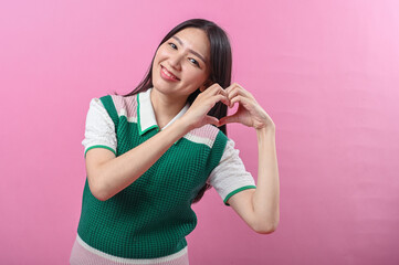Asian woman smiling and tilting her head while making a heart shape with both hands, wearing a...