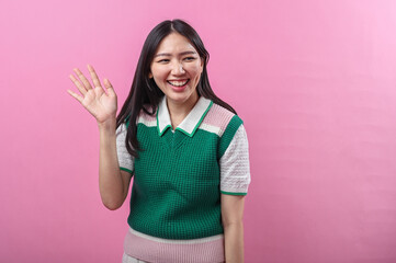 Asian woman waving and smiling in a friendly gesture, wearing a green and pink sweater with short...