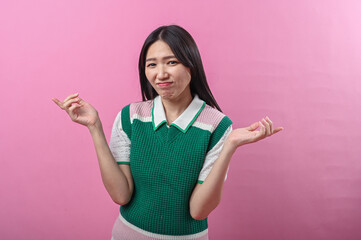 Asian woman in a green sweater vest making a confused shrugging gesture with both hands raised,...