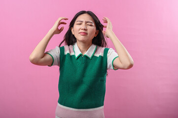 Fototapeta premium Asian woman pulling her hair in frustration and closing her eyes tightly, expressing extreme stress or anxiety, standing against a plain pink background