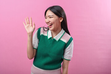 Asian woman waving and smiling in a friendly gesture, wearing a green and pink sweater with short...