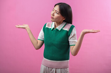 Asian woman shrugging with confused expression, wearing green and pink sweater vest, arms raised,...