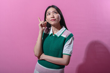 Asian woman smiling and pointing up with one finger, wearing green and pink sweater vest, standing...