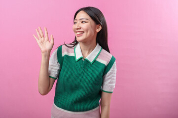 Asian woman waving and smiling in a friendly gesture, wearing a green and pink sweater with short...