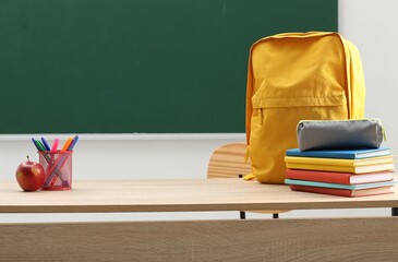 Different school stationery, backpack and red apple on wooden table near blackboard indoors