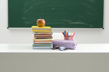Different school stationery, glasses and apple on white table near blackboard indoors