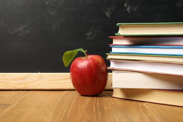 Different books and red apple on wooden table near blackboard, closeup. Space for text