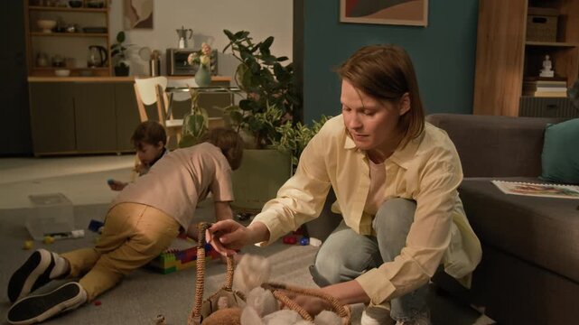 Tilt up shot of woman gathering up scattered plush toys putting them in basket bag and crouching down next to two sons who playing with blocks on floor