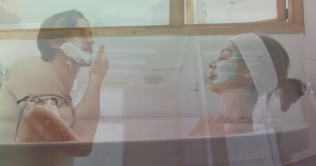 Shirtless man and woman wearing spa headband and facial mask grooming in bathtub, with shaving foam
