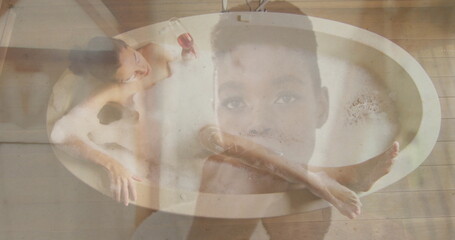 Reclining woman soaking in oval bathtub on plank floor, with foam, red wine glass, double exposure