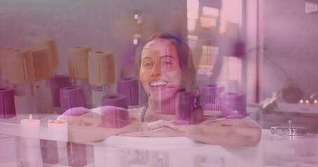 Leaning woman wearing swimsuit top in bathtub in spa room, with candles, towels, window, plant