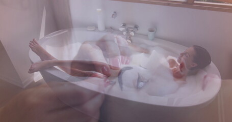 Reclining woman with hair pulled back soaking in bathtub in home bathroom, foam, smartphone overlay