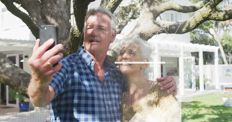 Taking selfie, sweater-clad woman and plaid-shirt man under oak tree in backyard, with smartphone