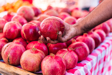 A close-up of a person's hand selecting a vibrant red pomegranate from a neatly arranged fruit...