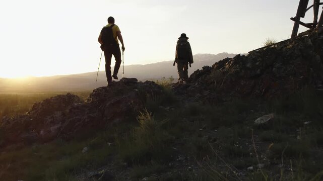 Two young active hikers with backpacks cross a mountain pass and begin their descent into a picturesque valley at sunset. Exploration, adventure, overcoming difficulties in the wild nature