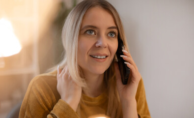 Smiling woman in a mustard sweater talking on the phone indoors. Bright natural light and soft...