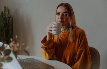 Thoughtful woman in mustard sweater holding a white mug while working on a laptop at home. Cozy...