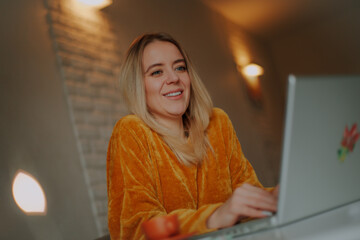 Cheerful woman in mustard sweater smiling while working on a laptop at home. Cozy indoor setting...