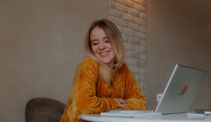 Smiling young woman in a mustard sweater sits at a desk with a laptop, turning her head with joy...