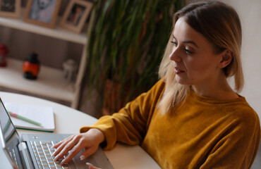 Focused young woman in a mustard sweater working on a laptop at home, seated at a white table with...
