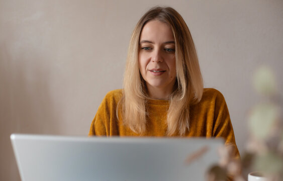Young woman in a mustard sweater working on a laptop at home. Focused expression, casual setting, soft daylight, and neutral background. Remote work or online learning concept.