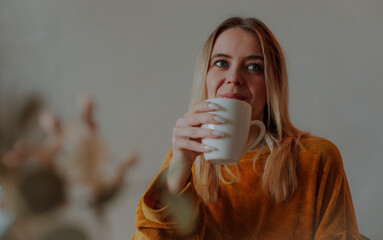 Young woman in a mustard sweater looking away while drinking from a white mug. Calm and cozy home...