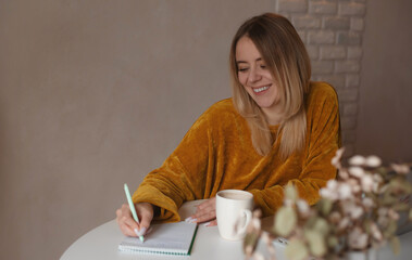 Smiling young woman in mustard sweater writing in a notebook at a white table with a cup of coffee....