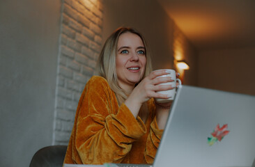 Young woman in a mustard sweater holding a mug while sitting in front of a laptop at home. Warm...