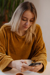 Young woman in mustard sweater sitting at a table, looking down at her smartphone with a focused...