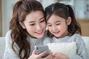 A Japanese woman and child watching a video on a smartphone. The mother, with a ponytail, is smiling at the phone screen while sitting next to the girl, who is holding a pillow in the living room.