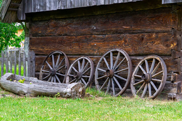 Obraz premium Wooden wagon wheels leaning against the wall of an old rural log barn. Traditional farming element set against green grass and a wooden fence