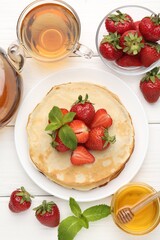 Delicious crepes with strawberries, honey, mint and tea on white wooden table, flat lay