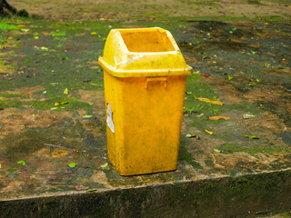 A yellow plastic trash bin stands alone on a damp, moss-covered concrete surface, surrounded by...
