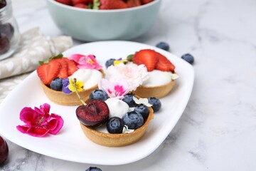 Sweet tartlets with berries and flowers on light marble table, closeup. Delicious dessert