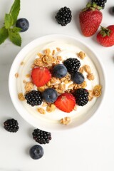 Tasty yogurt with granola and berries in bowl on white table, flat lay