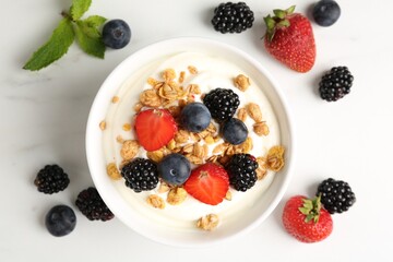 Tasty yogurt with granola and berries in bowl on white marble table, flat lay