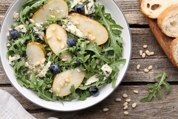 Tasty salad with arugula, pear, blue cheese, peanuts and blueberries in bowl on wooden table, flat lay
