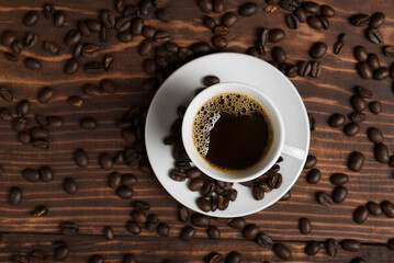 A cup of dark roasted coffee on a white saucer, surrounded by coffee beans on a brown wooden surface