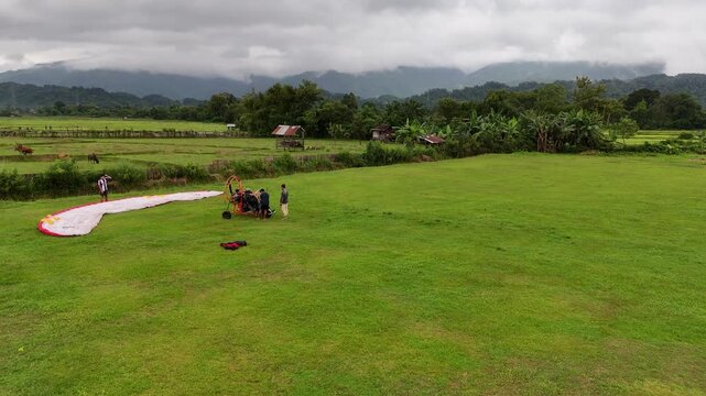Drone circles a paramotor during preflight in Vang Vieng, Laos, then follows as it takes off, with lush rice fields and scenic mountains in the background