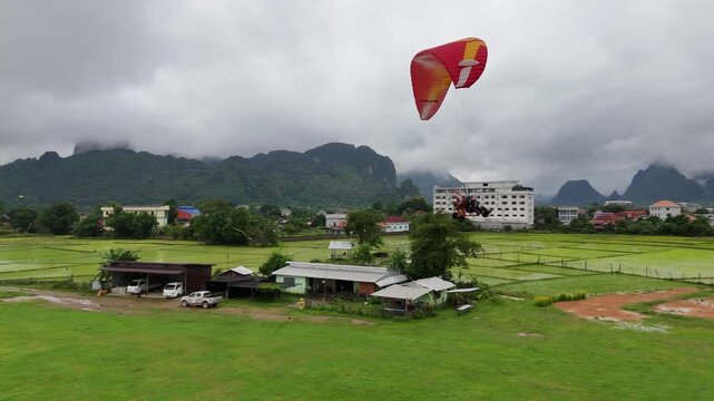 Drone follows a paramotor gliding over lush rice fields in Vang Vieng, Laos, ascending toward a dramatic mountain backdrop