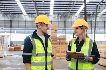 Smiling industrial workers discussing logistics details with clipboard inside organized pallet warehouse, surrounded by wooden materials for export and constructio