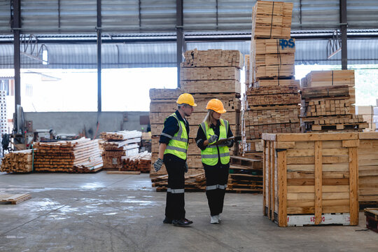 Male and female factory staff walking together through lumber storage area, discussing material management in organized industrial environment, captured in wide-angle shot.

