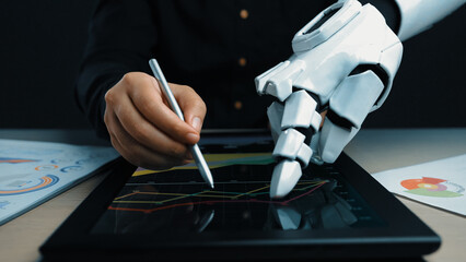 A human hand and a robotic hand cooperate in analyzing digital market data on a tablet, illustrating the synergy between technology and human insight in finance.Noogenesis